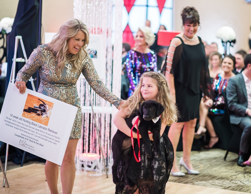 A trained black male hunting Labrador Retriever, named Mr. Black, was donated to the 2019 Vegas Gala live auction by Pam and Dr. David Black, Broadway Vet Clinic. Shown here with the puppy are Stacy Moore and Ryan Ragan holding Mr. Black, while Meredith Caddell, Foundation executive director from 2016 to 2020, looks on. Don’t miss out on the 20th anniversary Gala celebration! Get your tickets at Gala2026.GiveSmart.com.